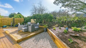A wooden deck with outdoor furniture, surrounded by a garden with various plants and a stone wall.