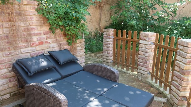 A wicker sofa with cushions against a brick wall, a wooden gate, and greenery in the background. - Müllers Wellness Ferienhaus (Döbrököz)