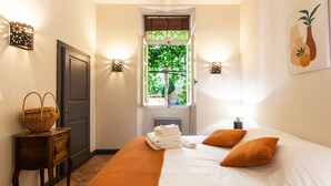 Un lit bien fait, avec une literie blanche et orange, une fenêtre donnant sur un paysage verdoyant et un luminaire fixé au mur.