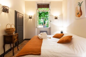 Een keurig opgemaakt bed met wit en oranje beddengoed, een raam met uitzicht op groen, en een wandlamp.