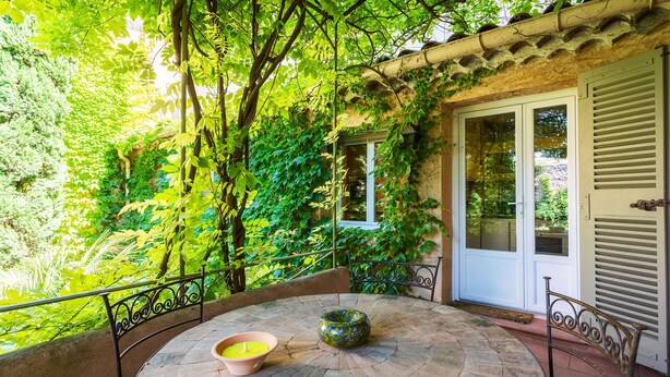 Une table ronde, surmontée d’un bol et d’une bougie, entourée d’une rambarde en métal, dans un jardin luxuriant agrémenté d’une porte et de volets.