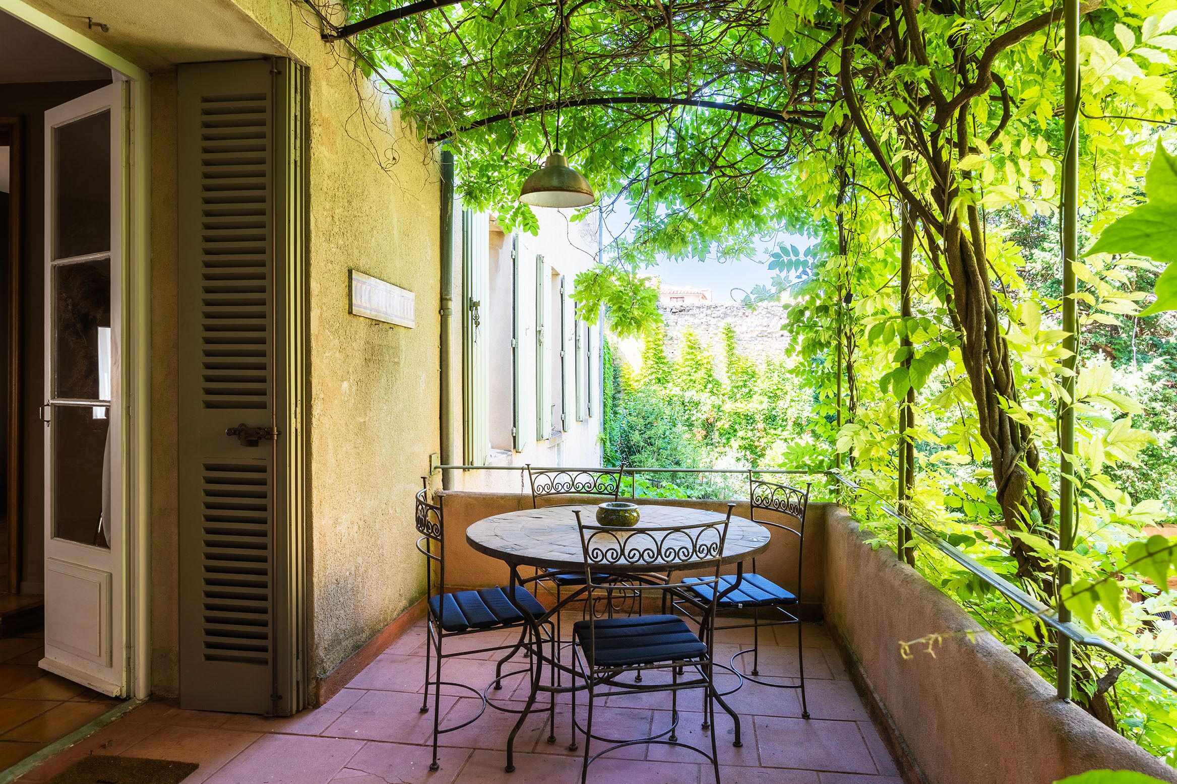 Ein Balkon mit rundem Tisch und Stühlen, umgeben von üppigem Grün und Blick auf ein Gebäude im Hintergrund.