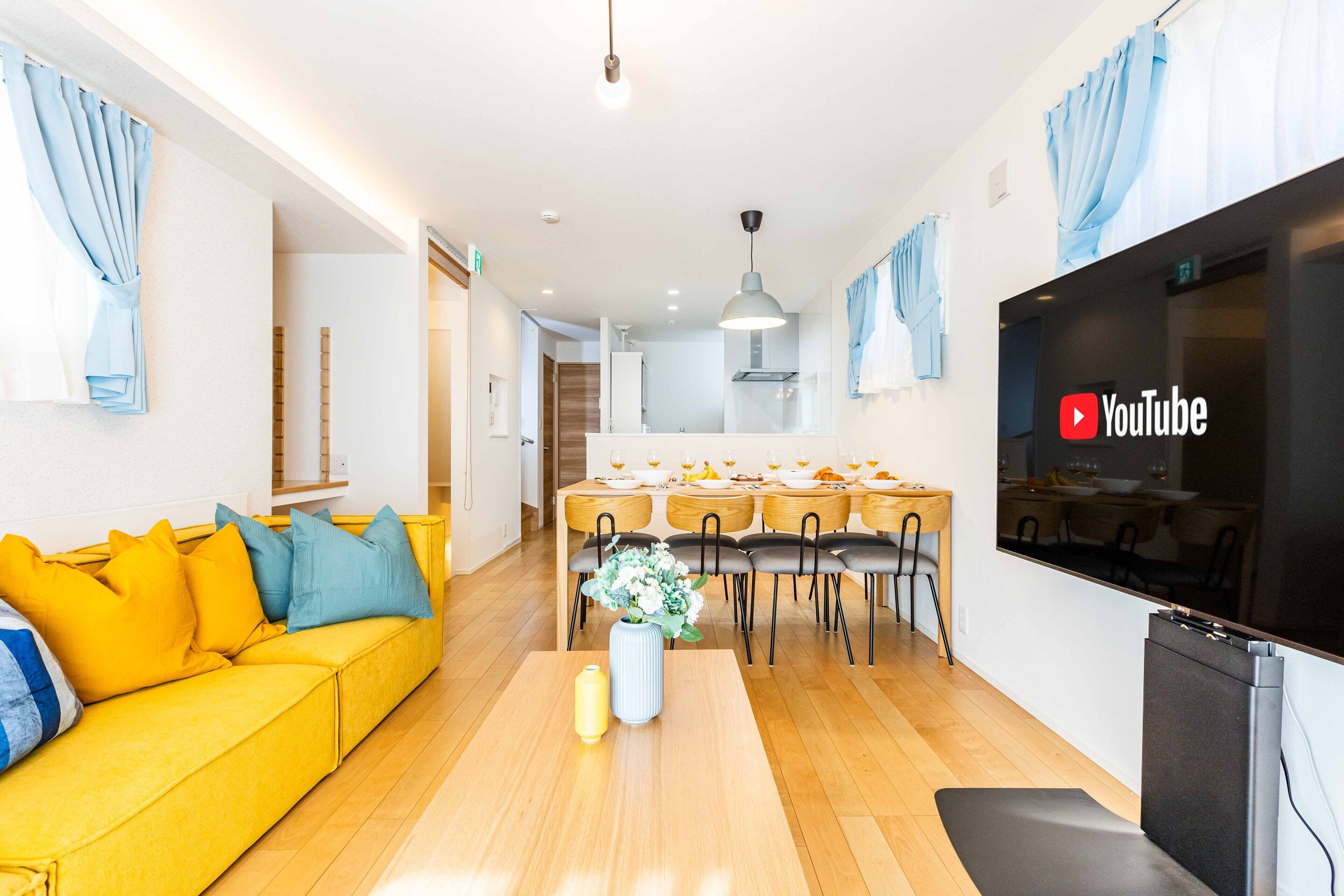 A modern living room with a yellow sofa, a dining area with a table set for four, and a flat-screen TV mounted on the wall.