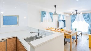 Una cucina moderna con isola centrale, zona pranzo con tavolo e sedie, e un'ampia finestra con tende azzurre.