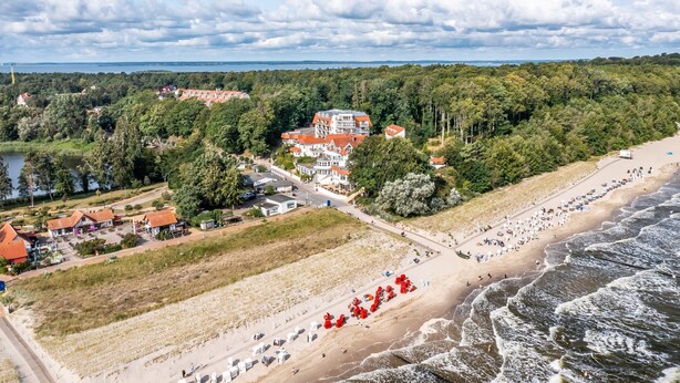 Vista aérea de una zona costera con playa, un edificio grande con tejados rojos y abundante vegetación.