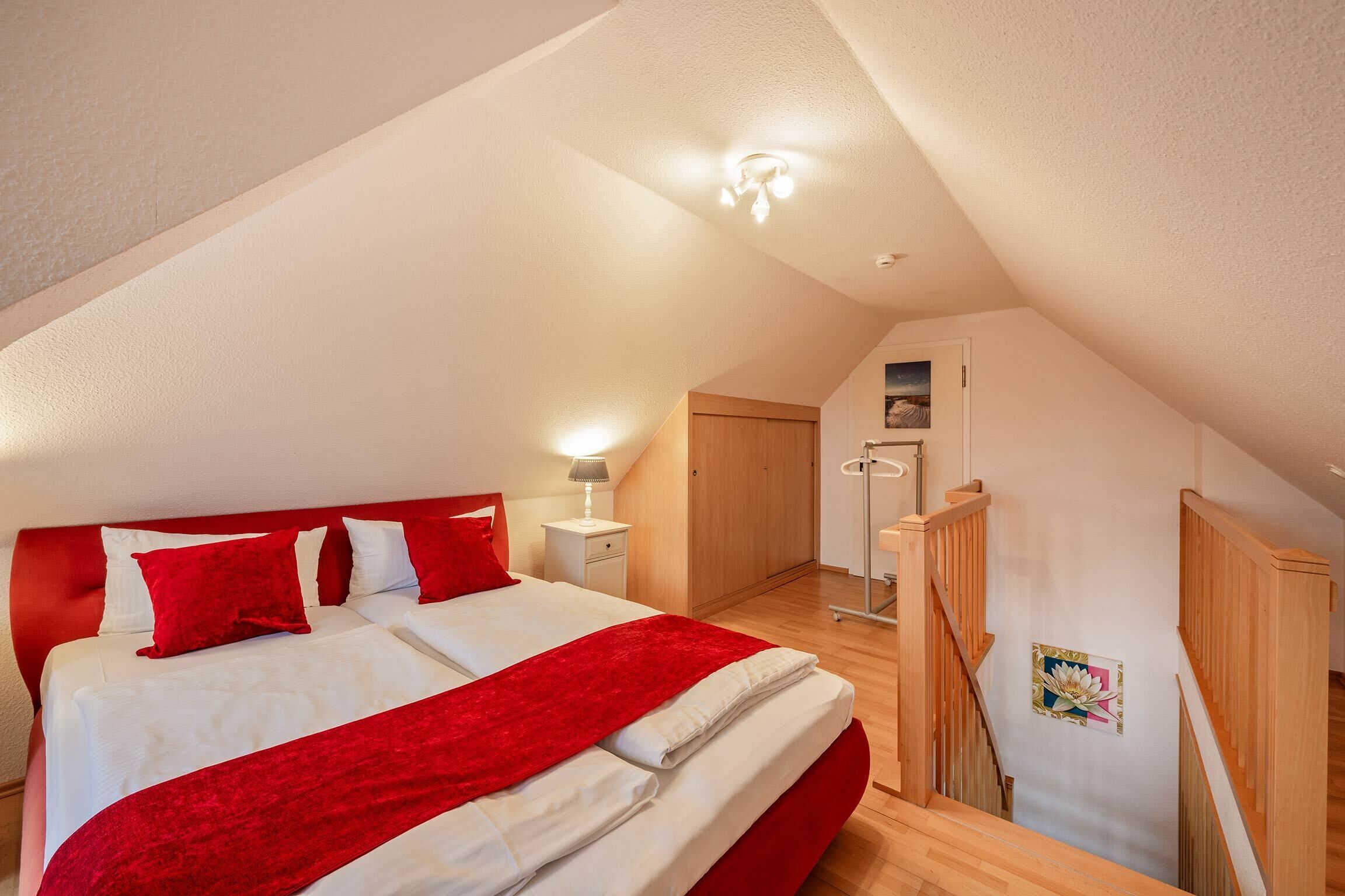 A bedroom with a large bed, red bedding, a wooden headboard, a nightstand with a lamp, and a wooden staircase.