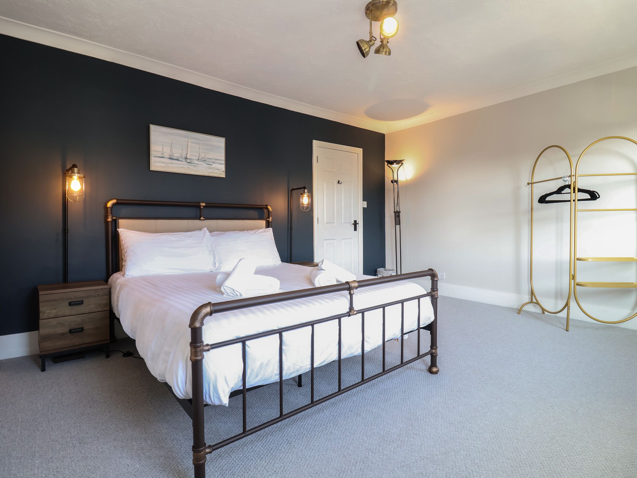 A neatly made bed in a room with a dark wall, a white door, and a gold coat rack.