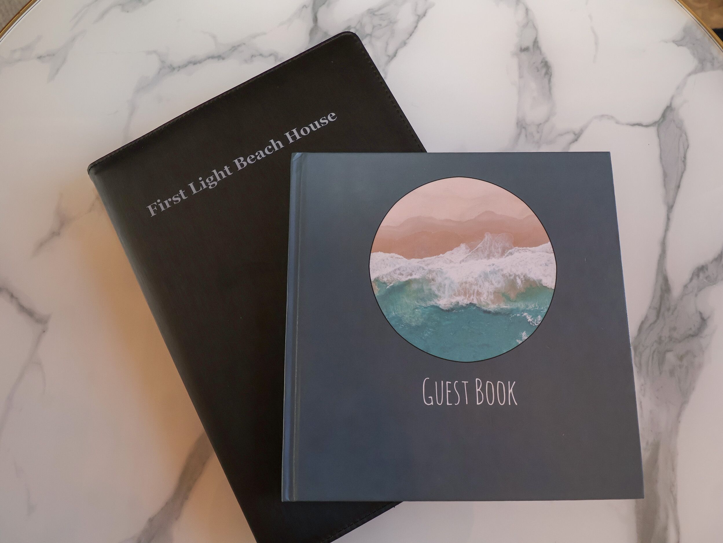 Two books on a marble surface. One book is titled "First Light Beach House" and the other is labeled "GUEST BOOK".