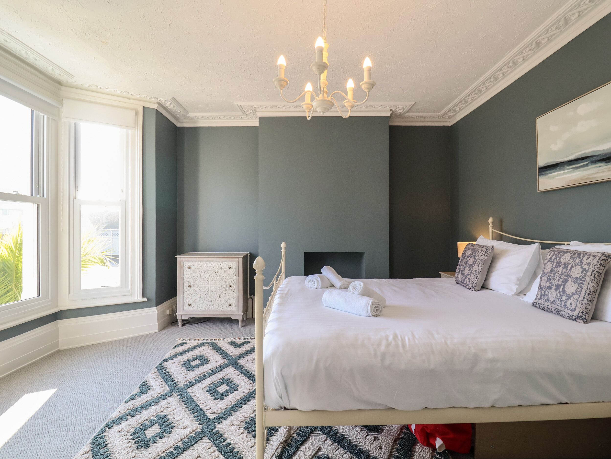 Un dormitorio con cama, mesita de noche, cómoda, candelabro y ventana con vista a follaje.