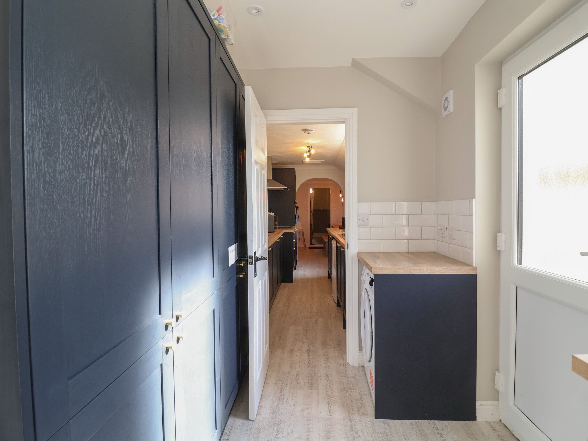 A narrow hallway with a dark door on the left, a light-colored tiled wall on the right, and a kitchen area in the background.
