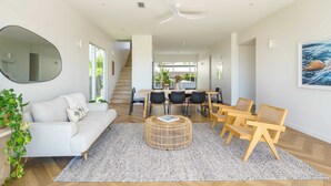 A modern living room with a sofa, coffee table, and chairs. A dining area with a table and chairs is visible in the background.