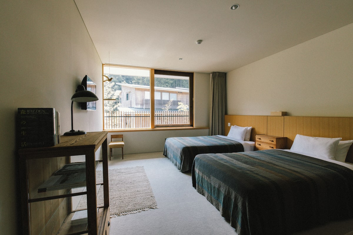 A hotel room with two beds, a desk, a chair, and a window with a view of buildings and trees.