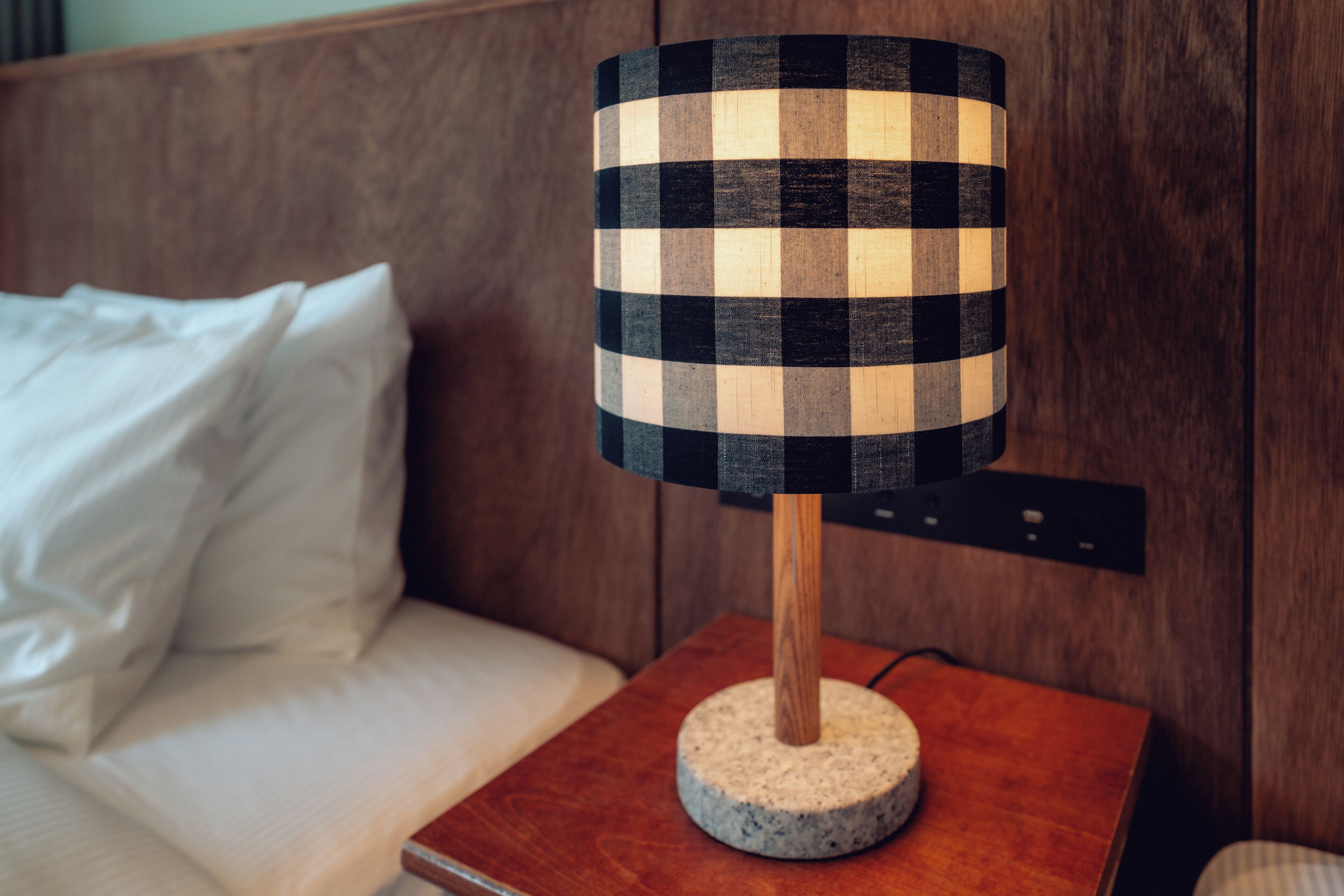 A checkered lamp on a wooden nightstand beside a bed with white bedding.