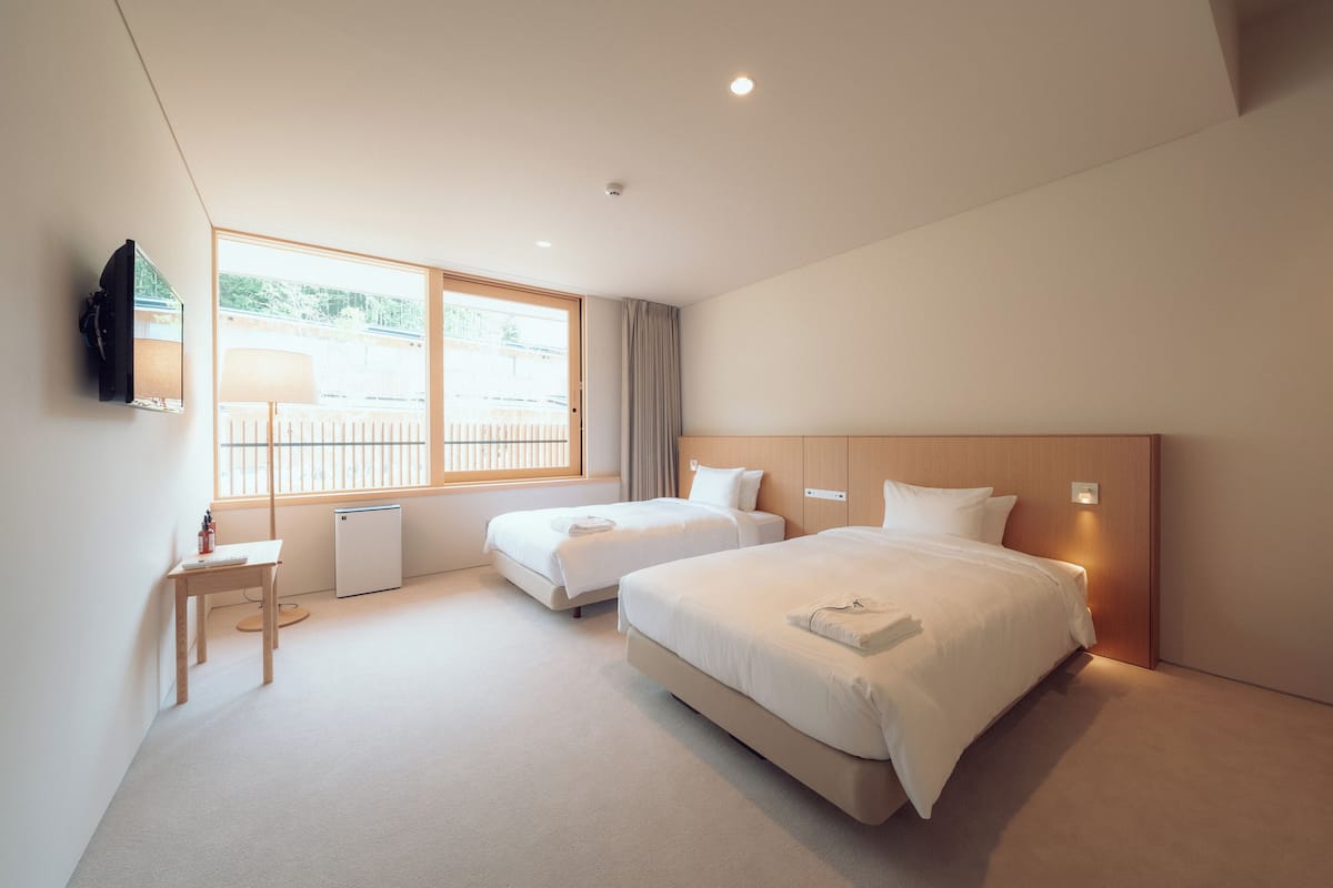 A hotel room with two beds, a desk, a TV mounted on the wall, and a window with a view of the outside.