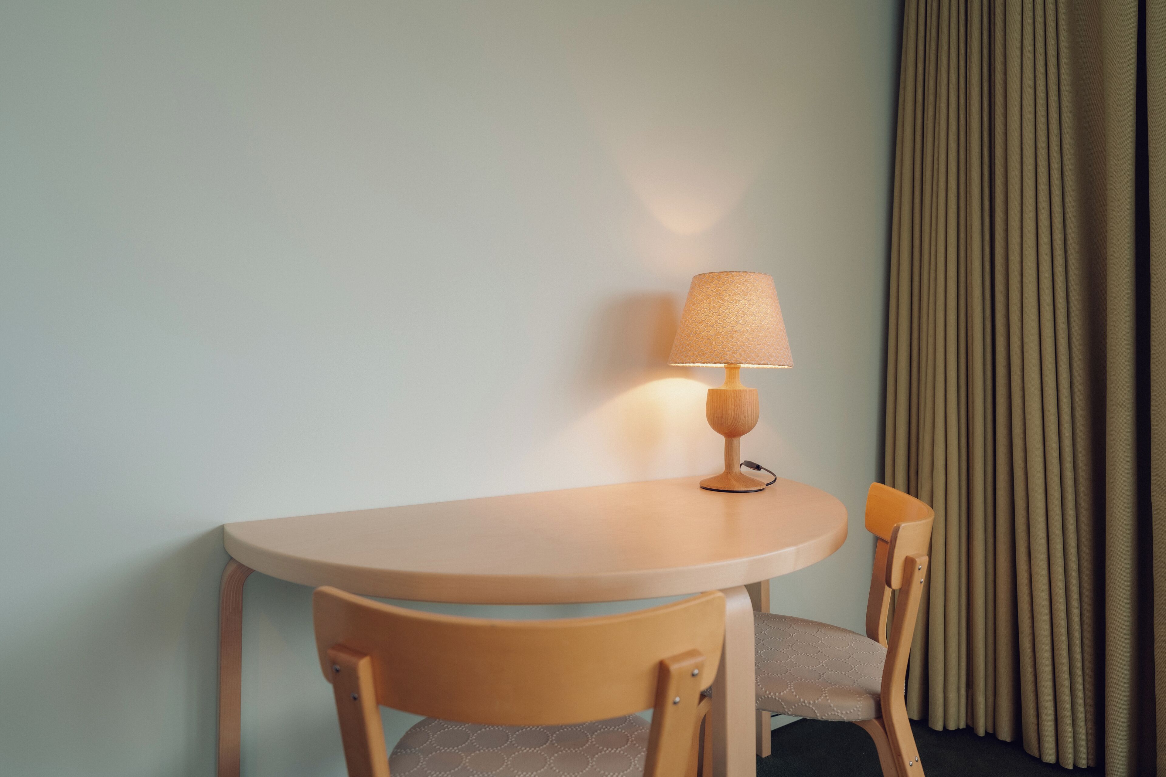 Et lille rundt bord med en lampe, en stol og et gardin i baggrunden.
