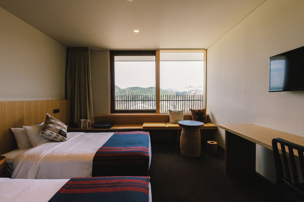 A hotel room with two beds, a desk, a chair, a small round table, a TV mounted on the wall, and a large window with a view of the outdoors.