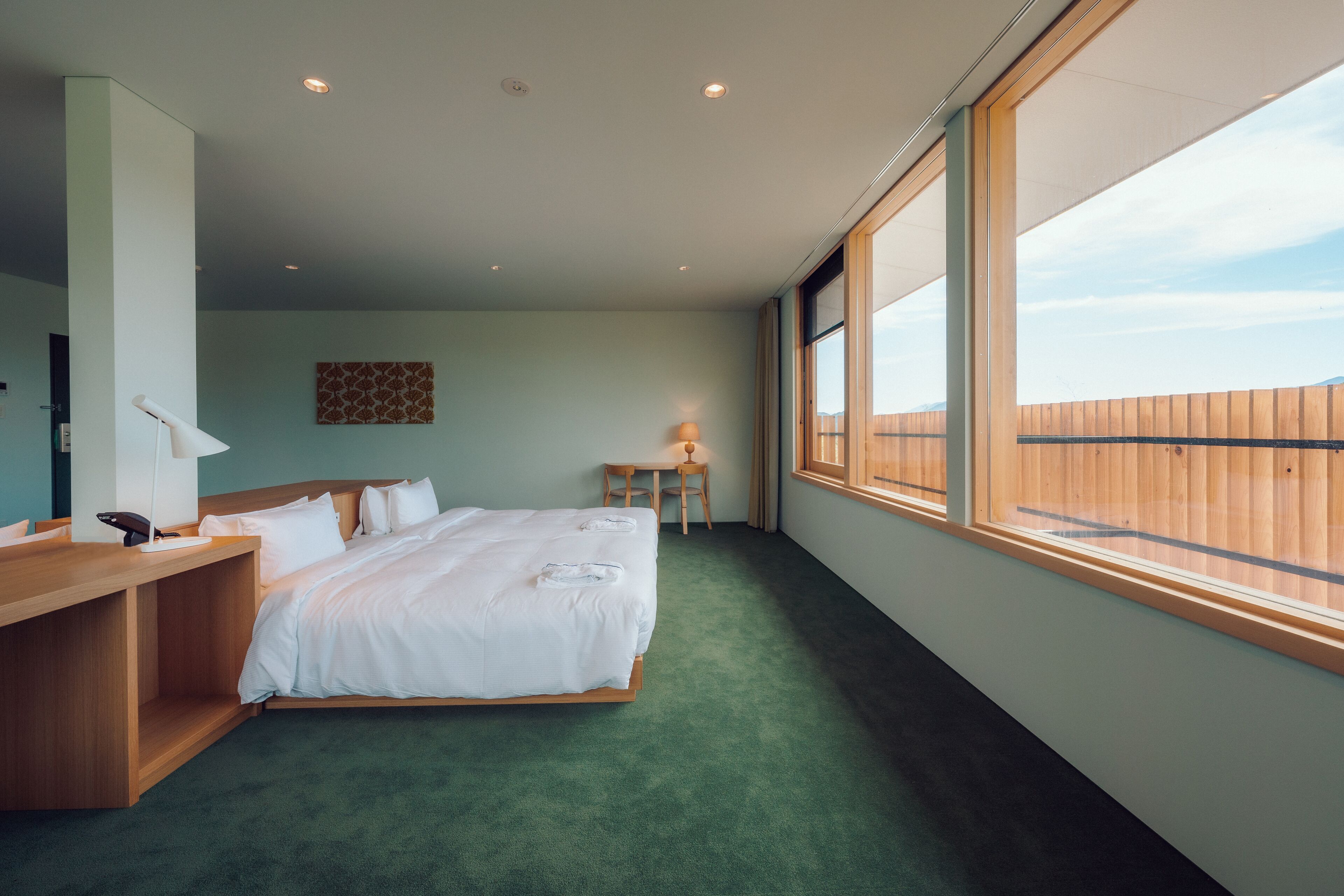 A hotel room with a large bed, a wooden desk, a small table, and a window with a view of a wooden fence.
