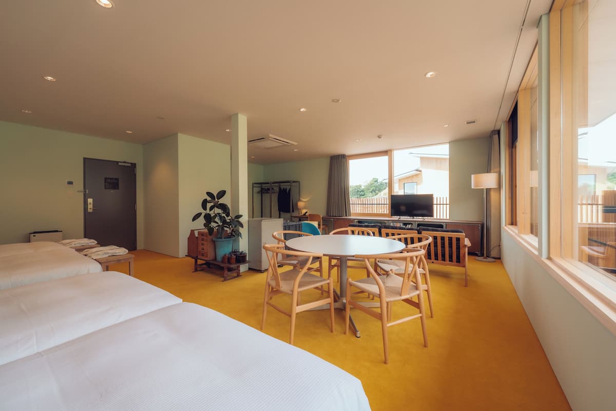 A hotel room with two beds, a dining area with a table and chairs, a TV, and a window with a view of the outside.
