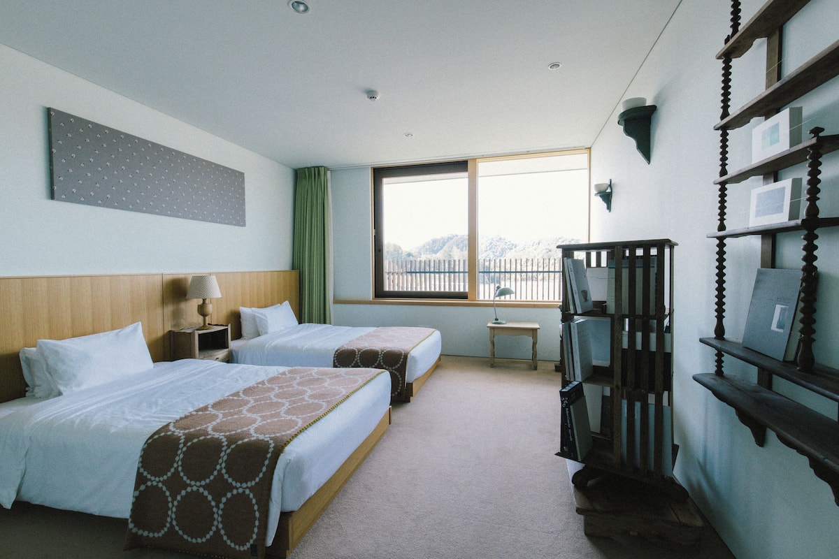 A hotel room with two beds, a large window, a wooden headboard, a small table, and a bookshelf.