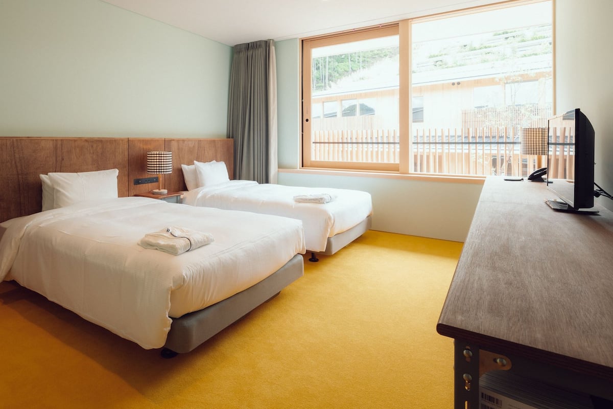 A hotel room with two beds, a desk, a lamp, and a window with a view of the outside.