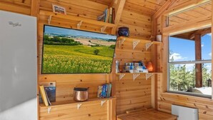 Interior de uma cabana de madeira com geladeira, televisão exibindo uma paisagem, prateleiras com livros e uma panela, e janela com vista para árvores.