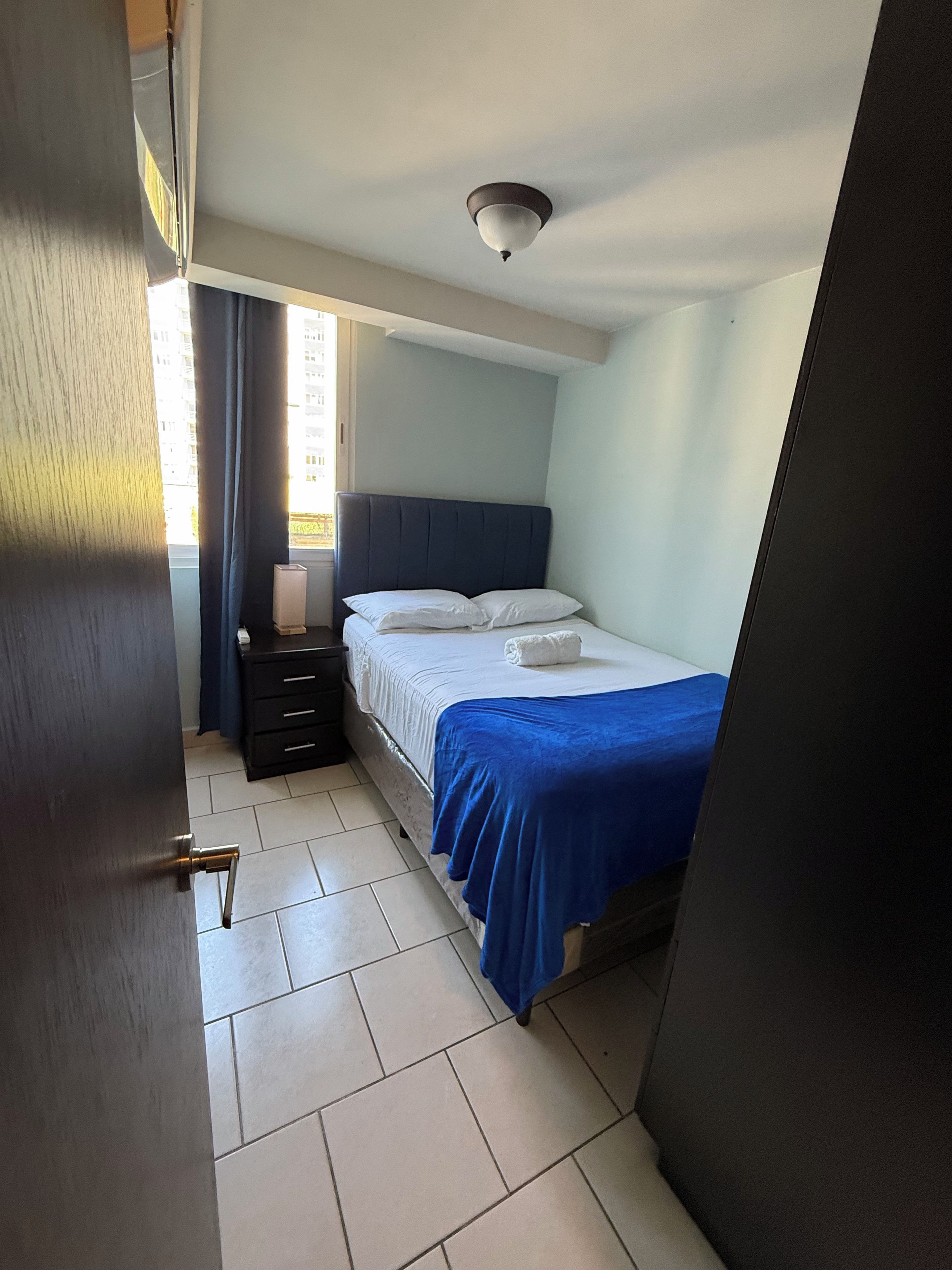 Une chambre à coucher comprenant un lit, une table de chevet, une commode, une fenêtre avec des rideaux et un luminaire au plafond.