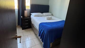 Une chambre à coucher comprenant un lit, une table de chevet, une commode, une fenêtre avec des rideaux et un luminaire au plafond.