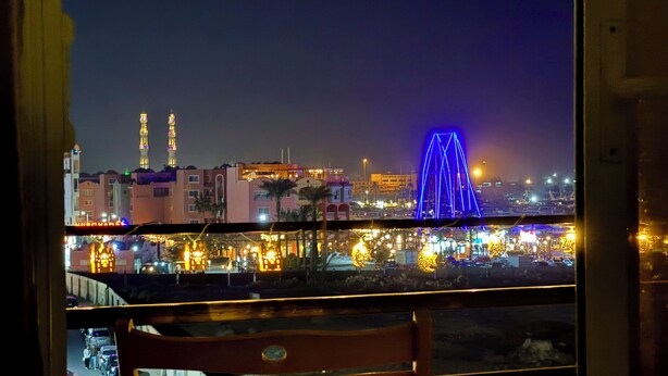 A view from a balcony with a chair, overlooking a cityscape at night with illuminated buildings and a prominent blue-lit bridge.