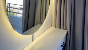 A room with a round mirror on a white console, a view of the sea and buildings, and a curtain on the right.