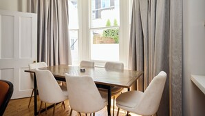 A dining area with a wooden table and four chairs, large windows with curtains, and a view of greenery outside.