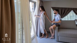 A room with a sofa, a large window with curtains, and a logo for 'ELIA Stone Villas'. - Premium Private Pool Villa, Elia Stone Villas (Thasos)