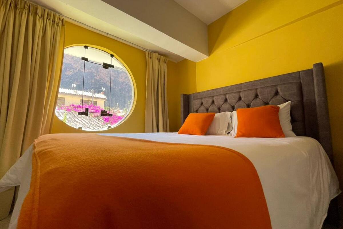 Quarto de hotel com uma cama grande, uma janela redonda com vista para um edifício e montanhas, e cortinas.