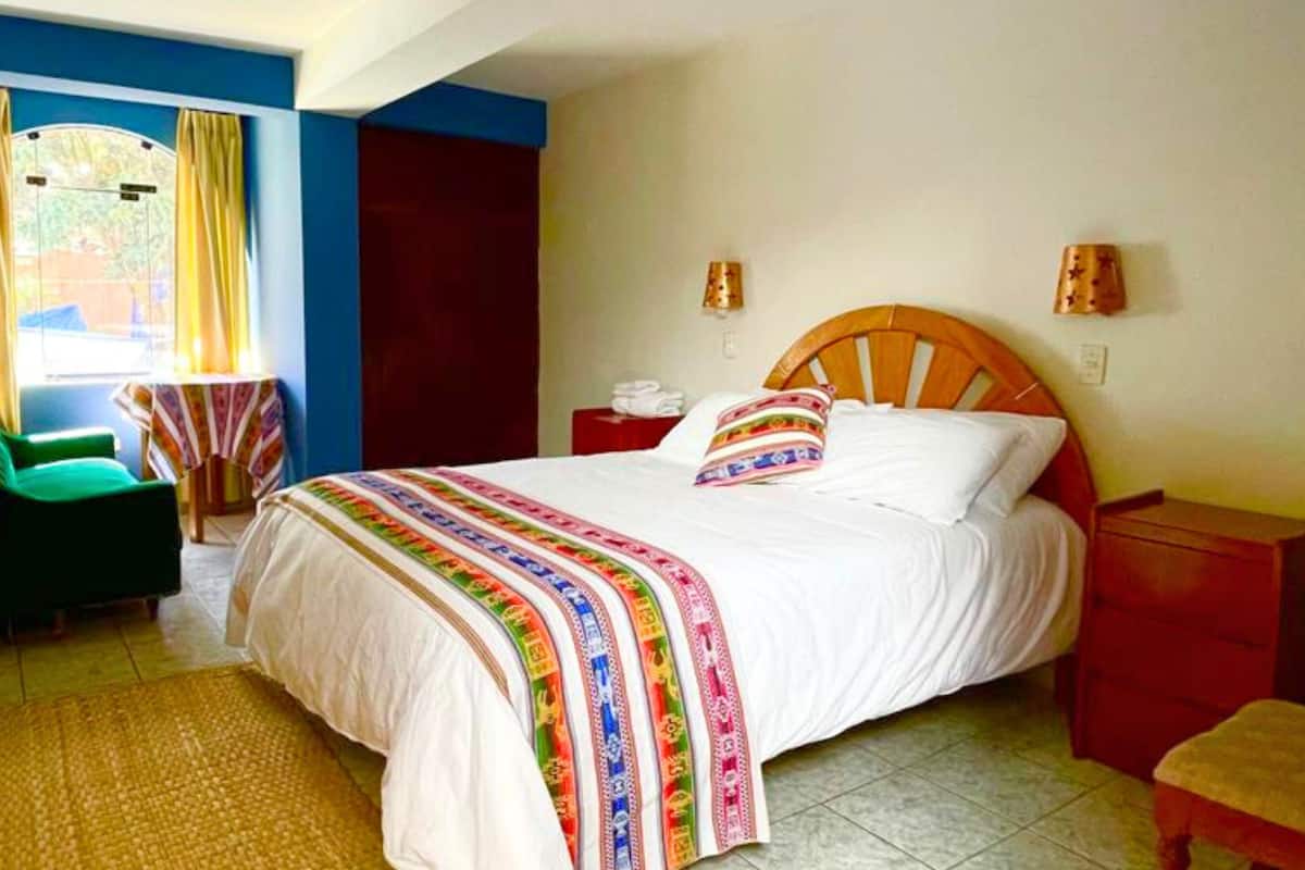 Quarto de hotel com cama, uma cadeira, uma mesinha e uma mesa de cabeceira.