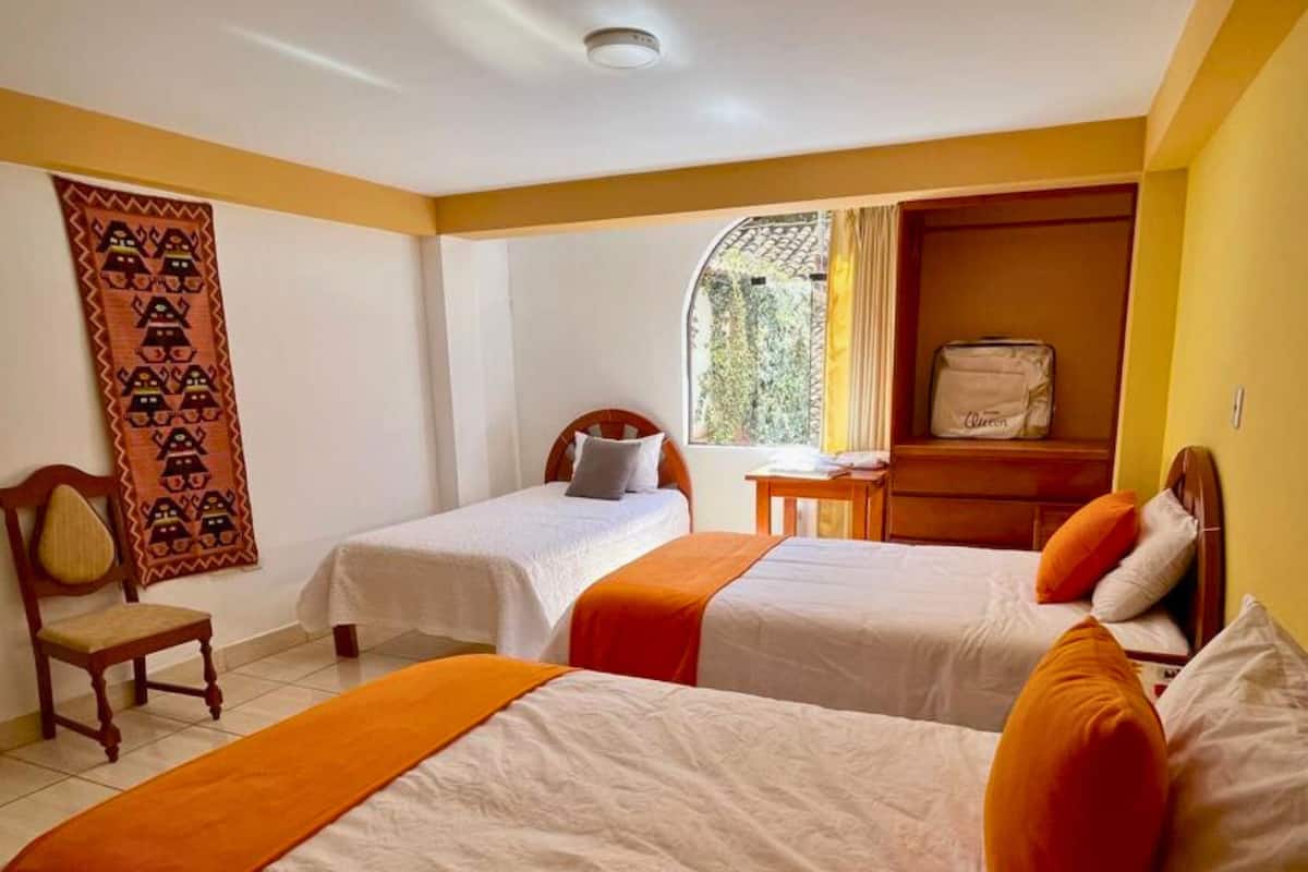 Quarto de hotel com duas camas de solteiro, uma cadeira de madeira, uma mesa pequena, uma mala e uma janela com cortinas.