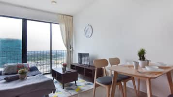 A modern living room with a sofa, a coffee table, a dining table with chairs, a TV, and a view of a cityscape through large windows.