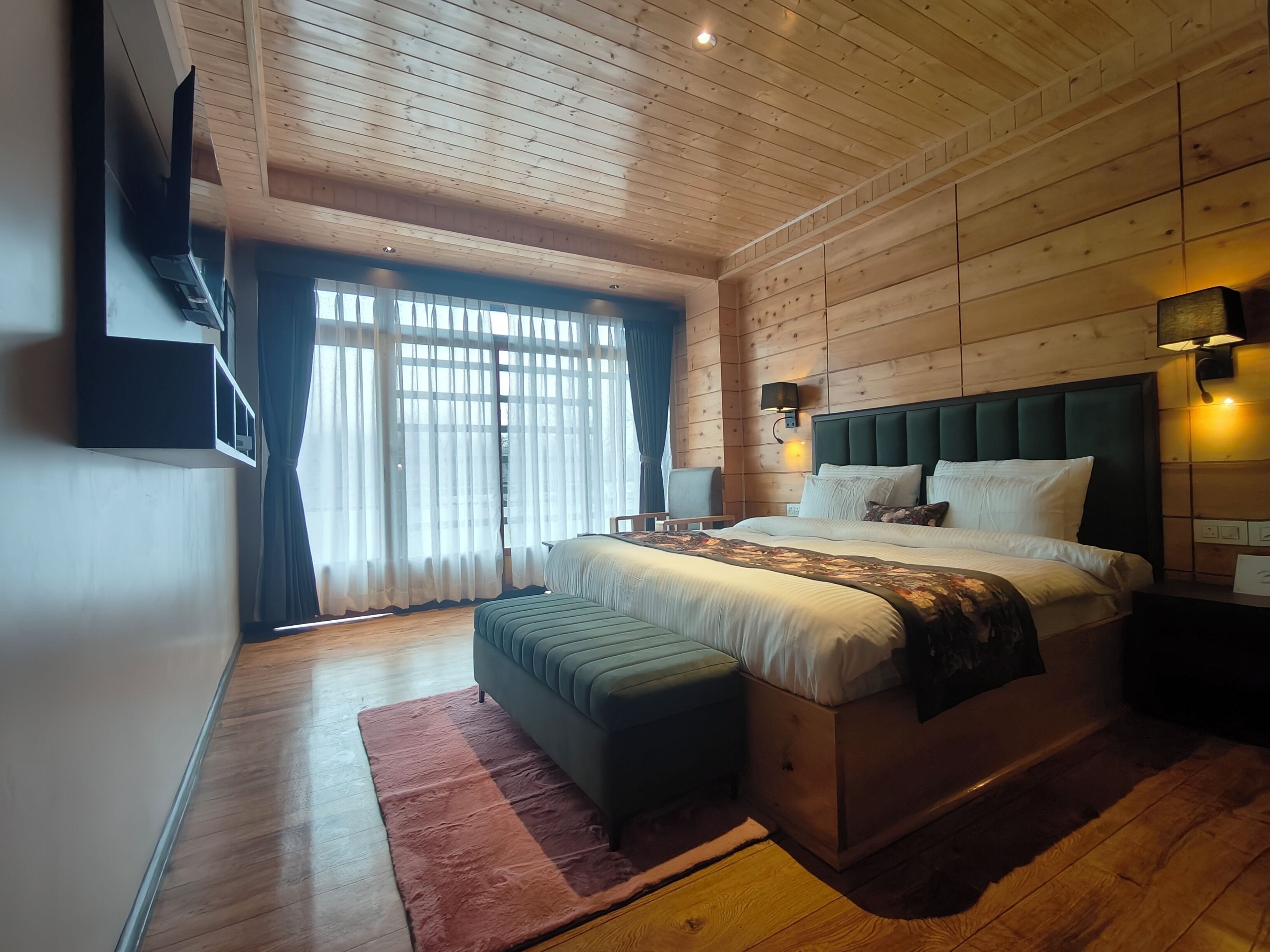 A bedroom with a large bed, a wooden ceiling, a TV mounted on the wall, and a window with sheer curtains.