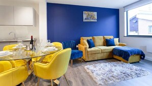 Un salon moderne avec une table à manger en verre, des chaises jaunes, un canapé beige agrémenté de coussins bleus et un tableau accroché au mur bleu.