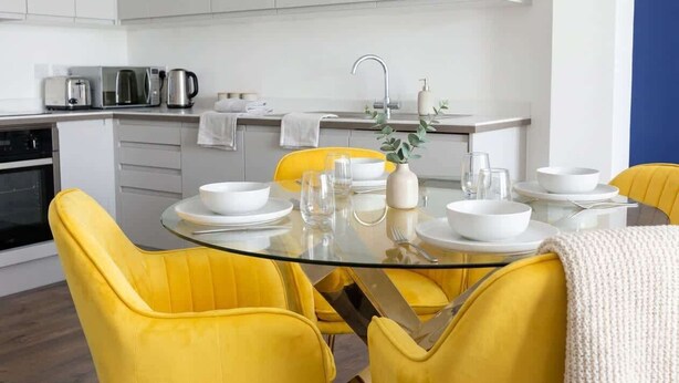 Une cuisine moderne avec des meubles blancs, une table à manger au plateau en verre et des chaises jaunes.