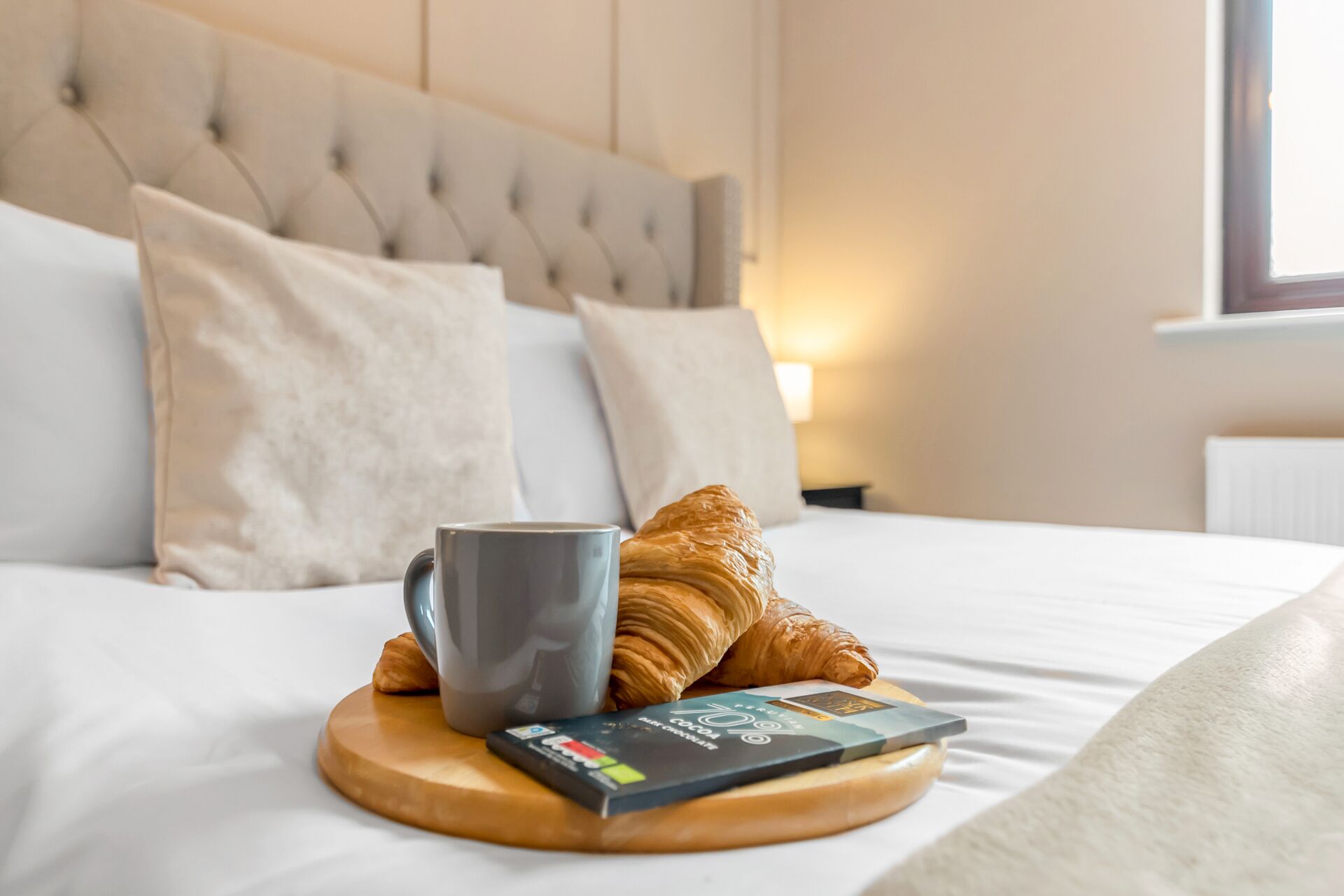 A bed with a grey mug, a croissant, and a chocolate bar on a wooden coaster.