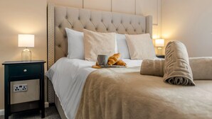 Une chambre d’hôtel avec une tête de lit capitonnée, un lit recouvert de draps blancs, deux tables de chevet avec des lampes et une couverture pliée posée sur le lit.