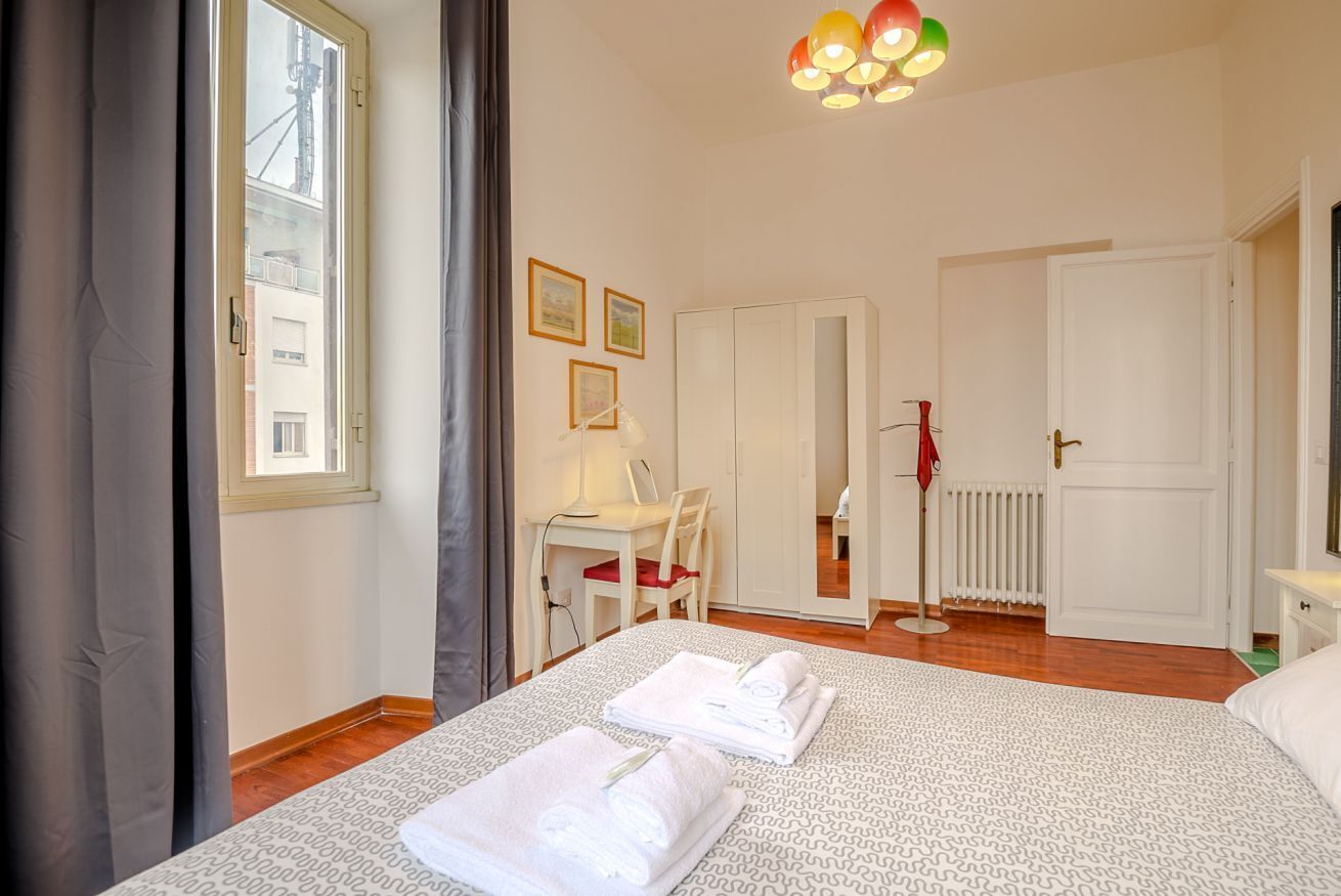 A hotel room with a bed, a desk, a chair, a radiator, and a window with curtains.