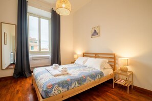 A bedroom with a wooden bed, a window with curtains, a wall-mounted lamp, and a small bedside table.