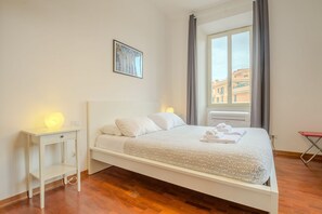 A single-bed room with a white headboard, a bedside table with a lamp, a window with curtains, and a painting on the wall. - Candia Cento Apartment Vatican Museums (Rome)
