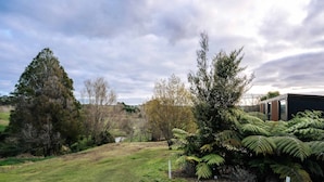 A grassy area with trees and ferns, a building with a modern design, and a cloudy sky. - Kiwi Farm Cabin 2 by Tiny Away (Waitoki)