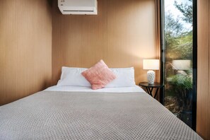 A neatly made bed with a white and pink pillow, a small bedside table with a lamp, and a view of greenery through a window.