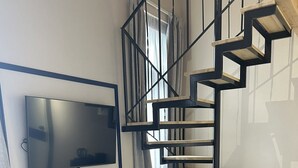 A modern living room with a black spiral staircase, a flat-screen TV mounted on the wall, a dark console table, and a potted plant. - Living Yard Thissio (Athens)