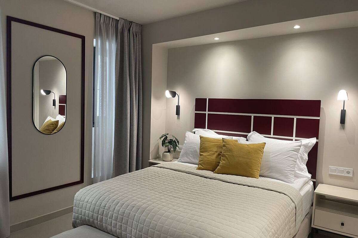 A modern bedroom with a large bed, a bedside table, a nightstand, a wall-mounted mirror, and a window with curtains.