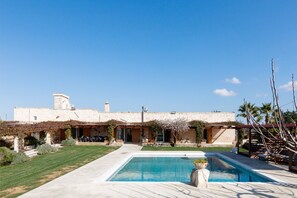 A pool area with a clear sky, a building with stone walls, and a garden with a pergola.