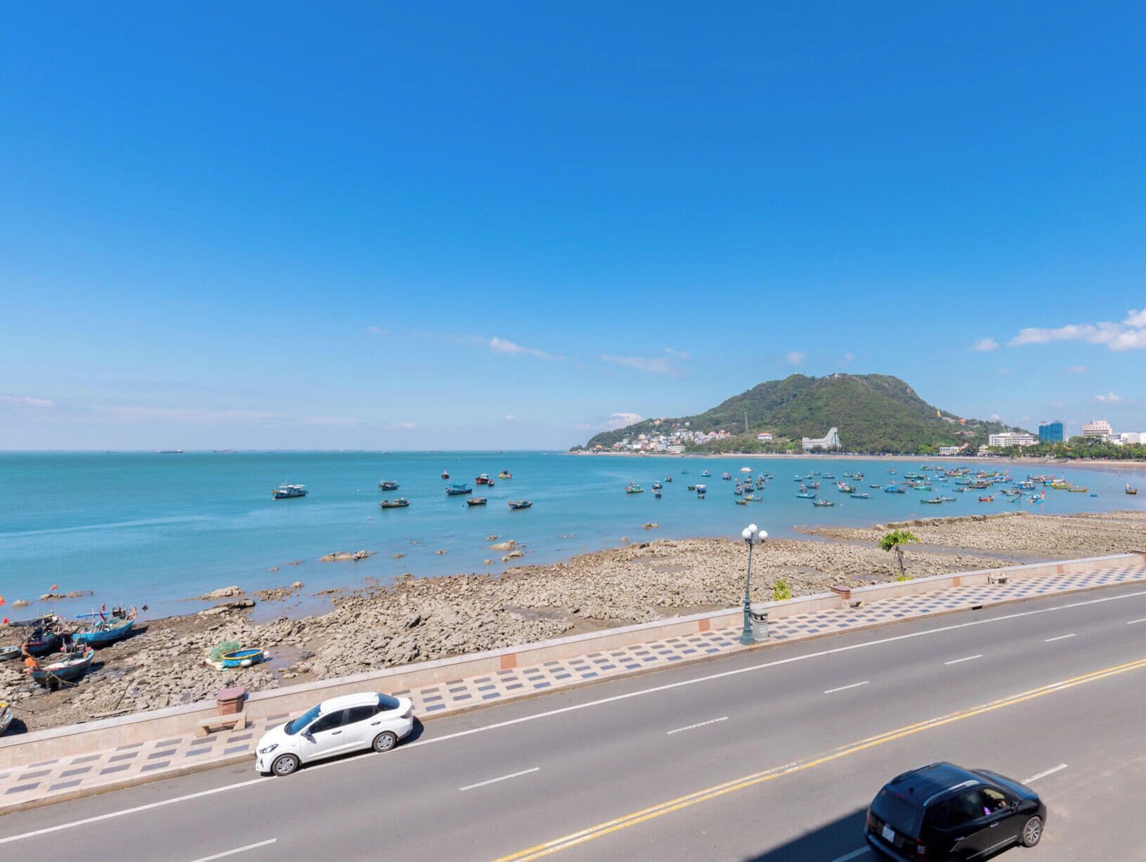A coastal road with parked cars, a beach with boats, and a hilly island in the background.