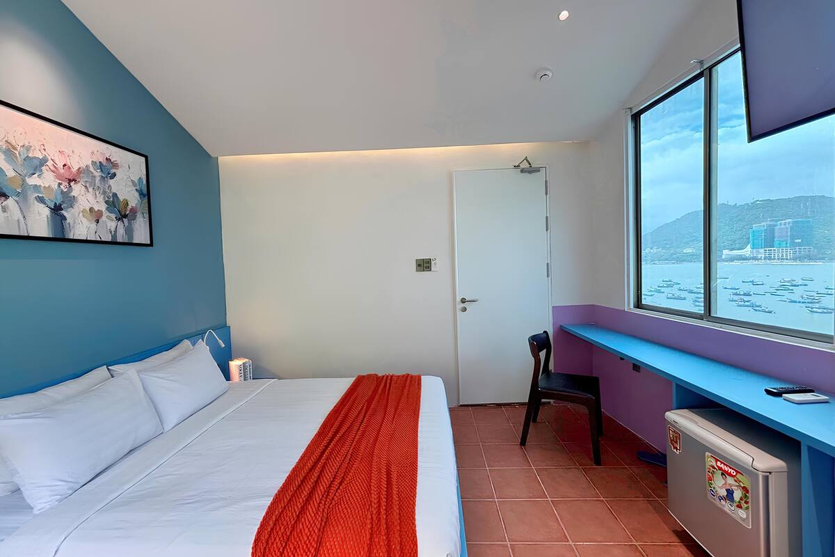 Un dormitorio con cama, escritorio, silla y ventana con vista a barcos.